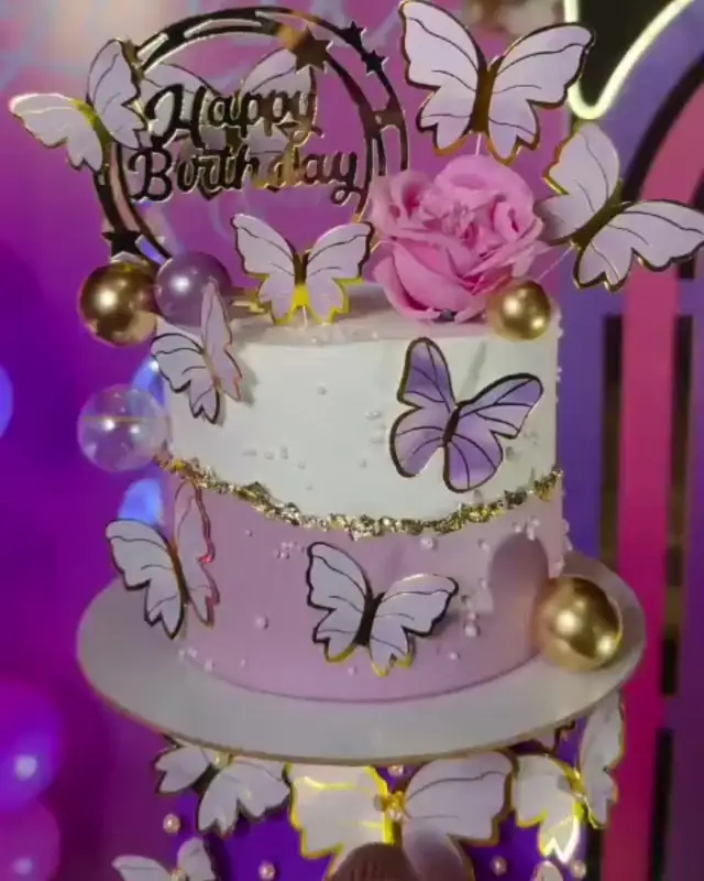 A two-tier white and lilac cake covered in paper butterflies, a fresh pink rose and gold pearl beads, with a 'Happy Birthday' topper.