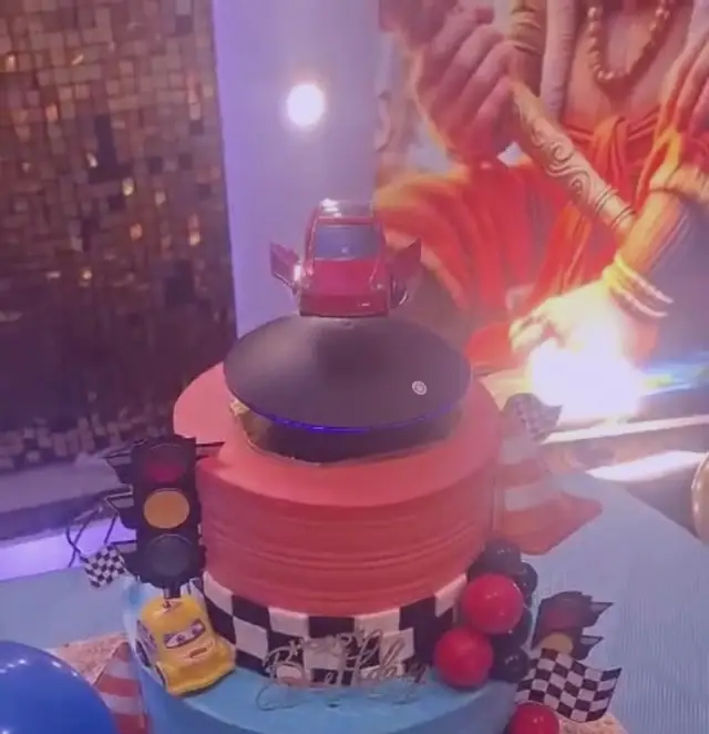 A two-tier orange and blue cake with a red toy car hovering above it on a hidden support, a traffic-light prop and a checkered ribbon at the base.